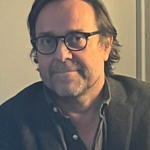 Headshot of a person with medium-length hair and glasses, wearing a dark button-up shirt and blazer, looking at the camera.